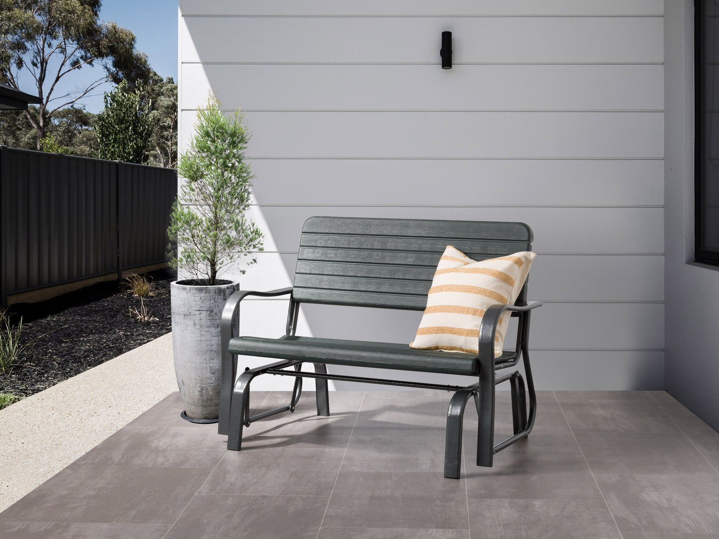 Gray metal gliding patio bench with curved armrests and slatted backrest, set on a wooden deck.